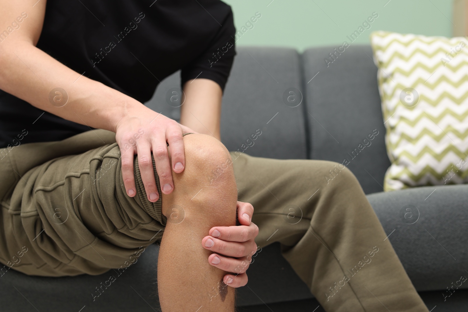 Photo of Young man suffering from pain in knee on sofa at home, closeup