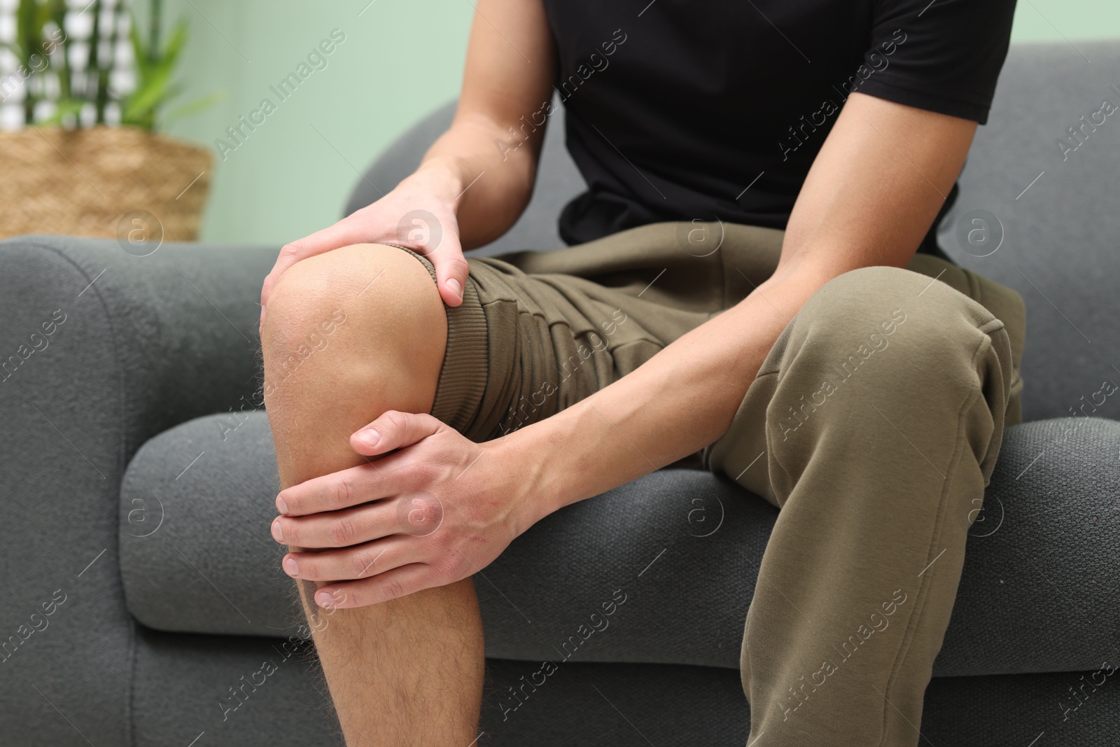 Photo of Young man suffering from pain in knee on sofa at home, closeup