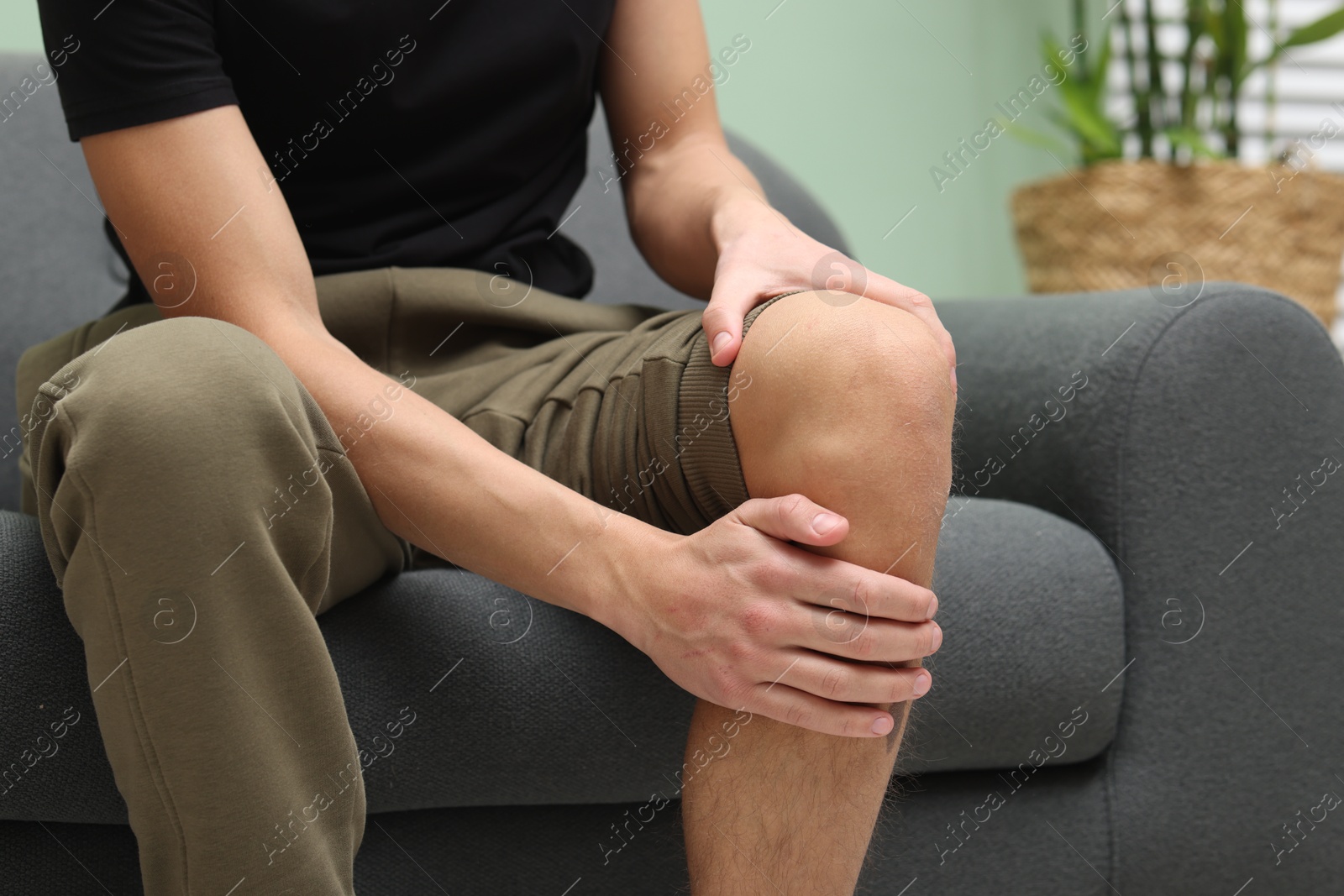 Photo of Young man suffering from pain in knee on sofa at home, closeup