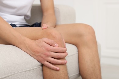 Photo of Young man suffering from pain in knee on sofa at home, closeup