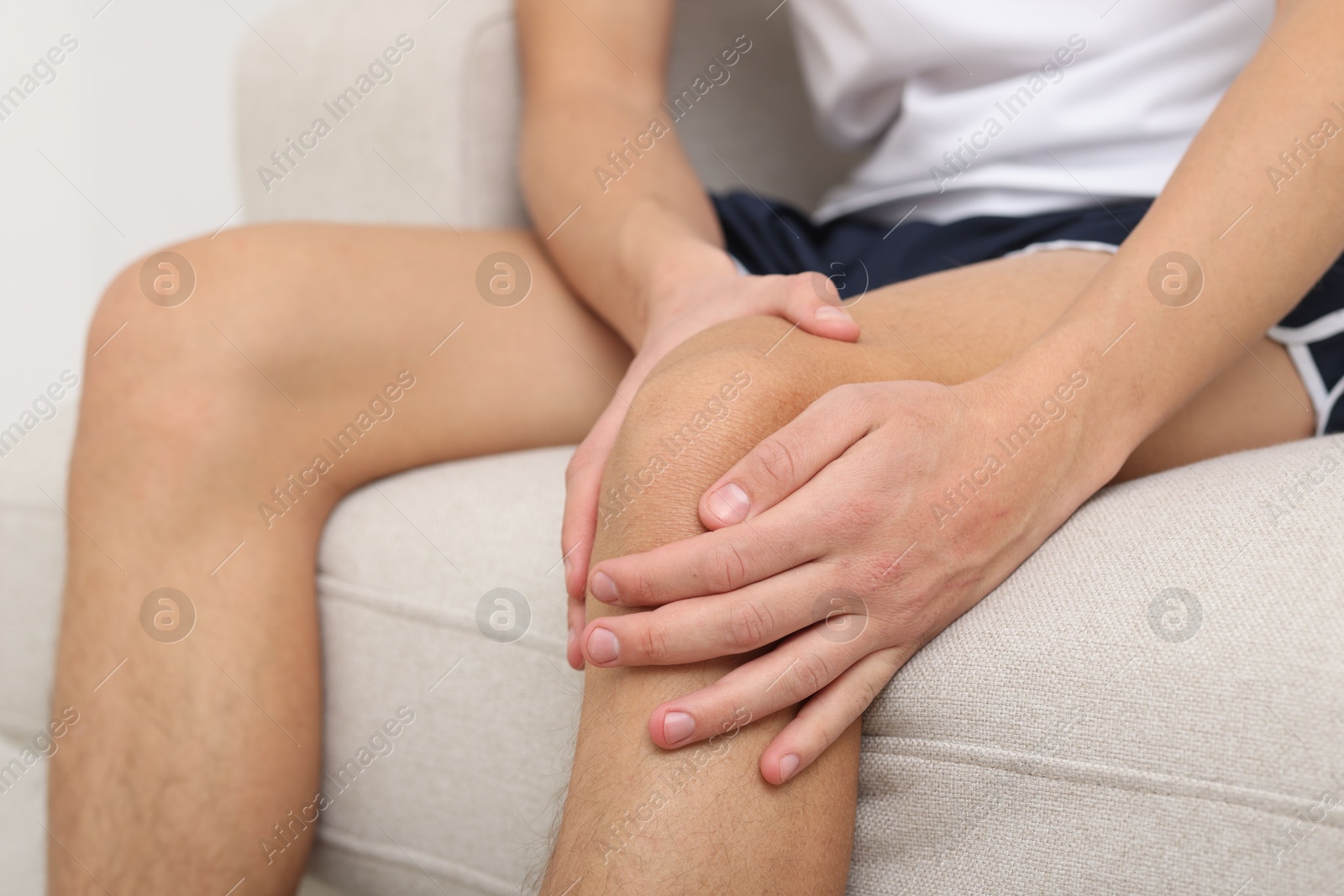 Photo of Young man suffering from pain in knee on sofa at home, closeup