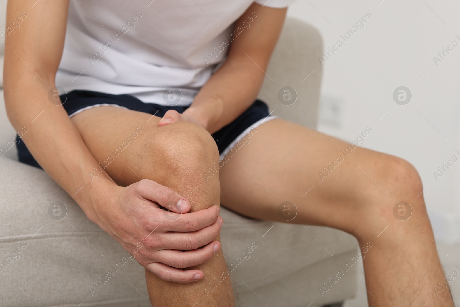 Photo of Young man suffering from pain in knee on sofa at home, closeup