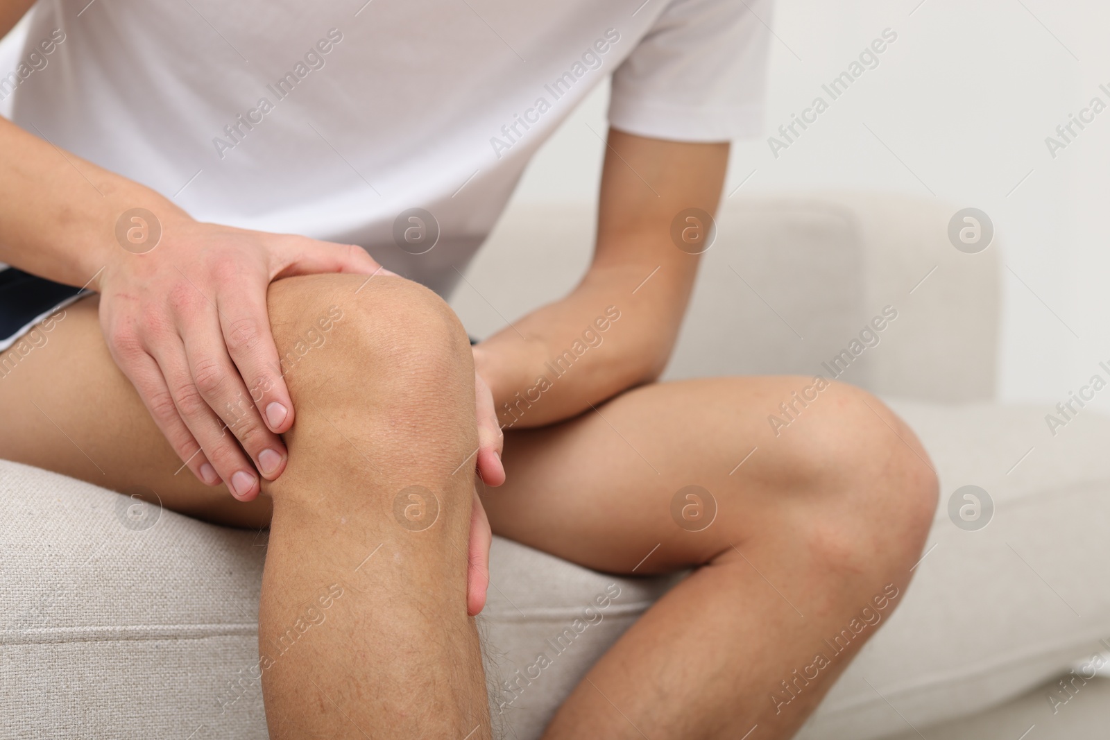 Photo of Young man suffering from pain in knee on sofa at home, closeup