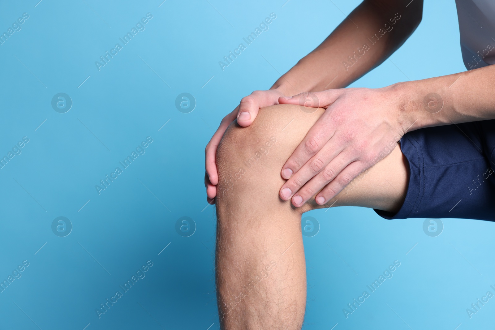 Photo of Young man suffering from pain in knee on light blue background, closeup. Space for text