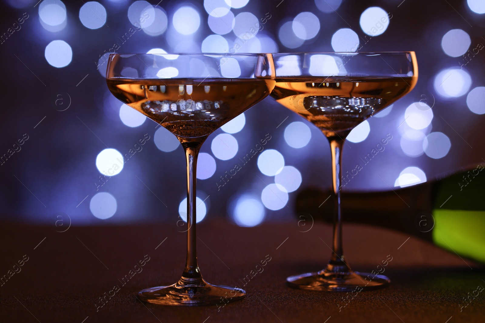 Photo of Champagne in glasses and bottle on table against dark background with blurred lights, closeup