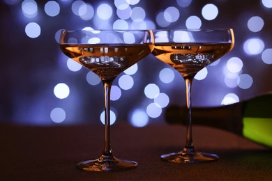 Photo of Champagne in glasses and bottle on table against dark background with blurred lights, closeup