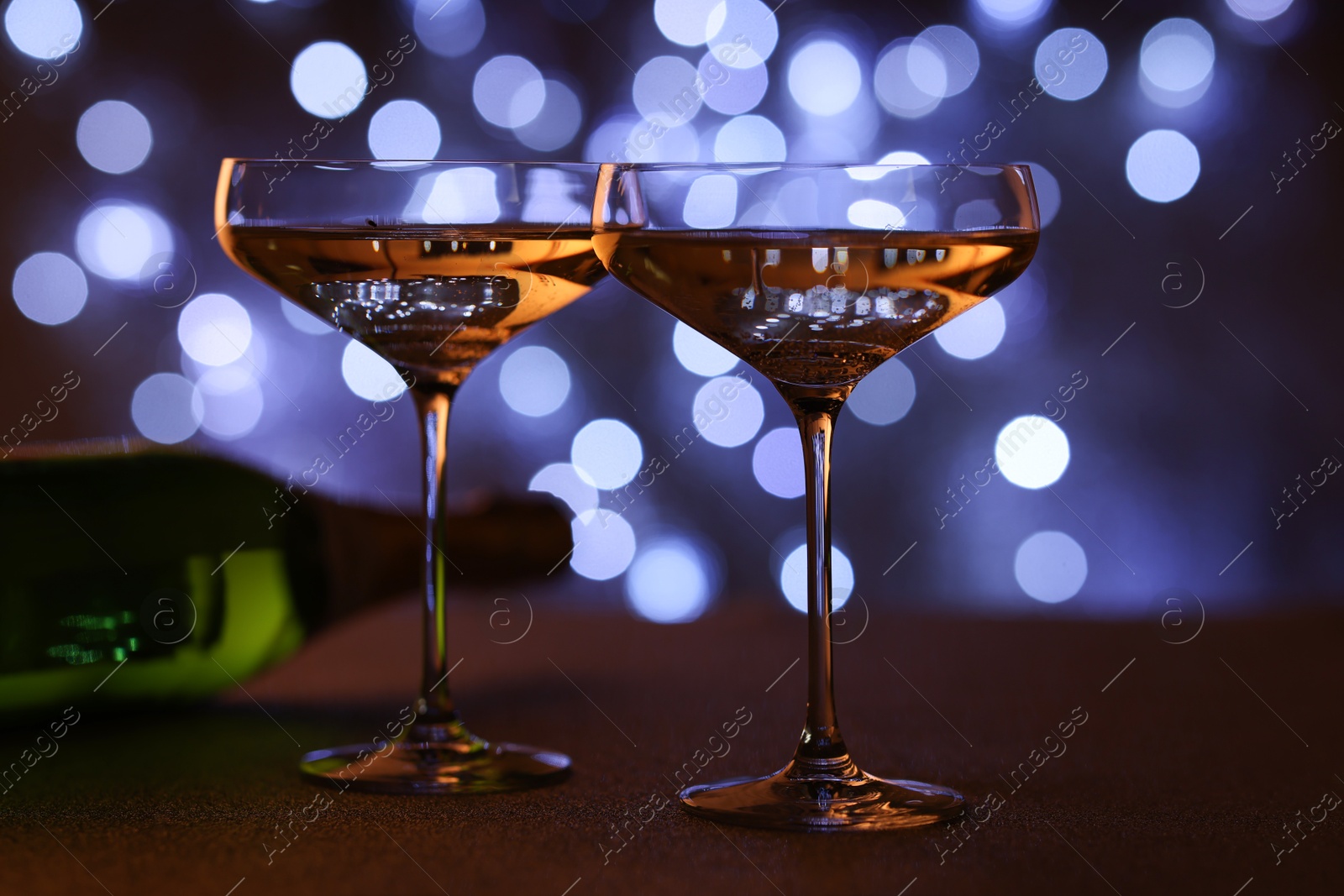 Photo of Champagne in glasses and bottle on table against dark background with blurred lights, closeup