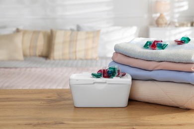 Photo of Detergent capsules in container and clean laundry on wooden table indoors. Space for text