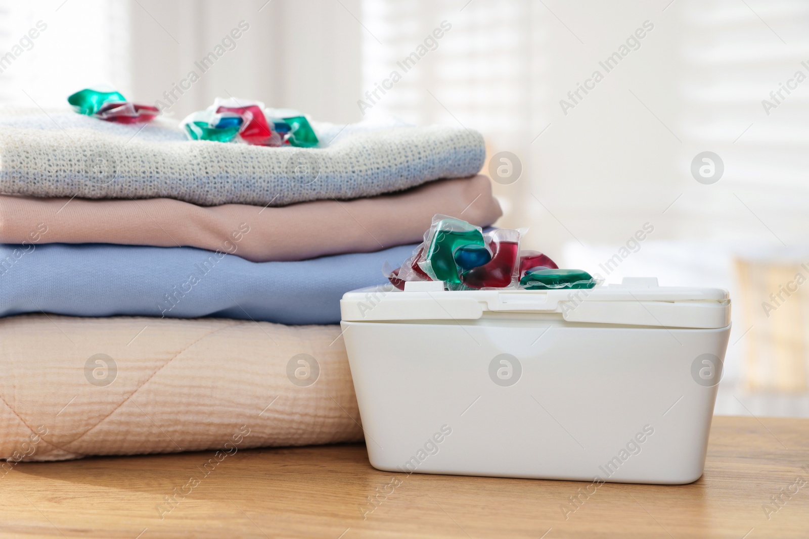 Photo of Detergent capsules in container and clean laundry on wooden table indoors