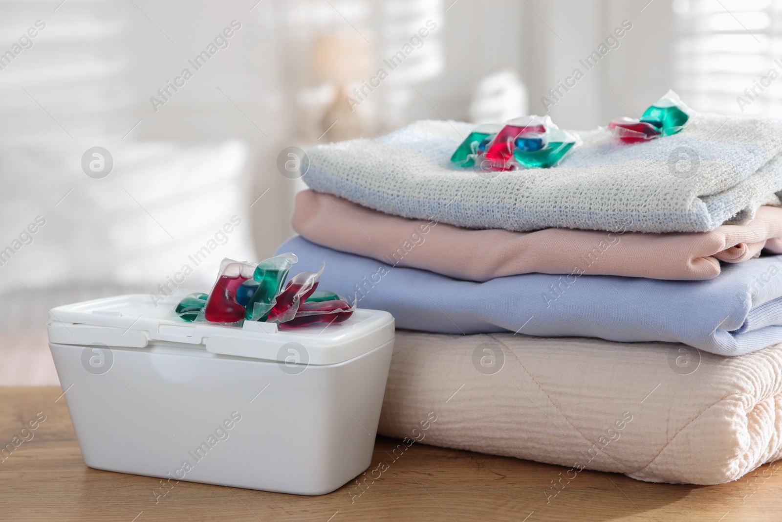 Photo of Detergent capsules in container and clean laundry on wooden table indoors