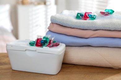 Photo of Detergent capsules in container and clean laundry on wooden table indoors