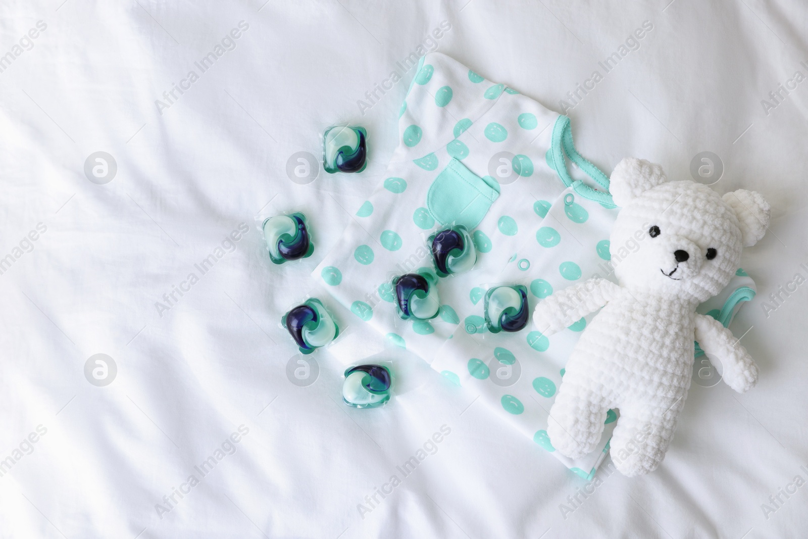 Photo of Laundry detergent capsules, teddy bear and baby onesie on white fabric, flat lay