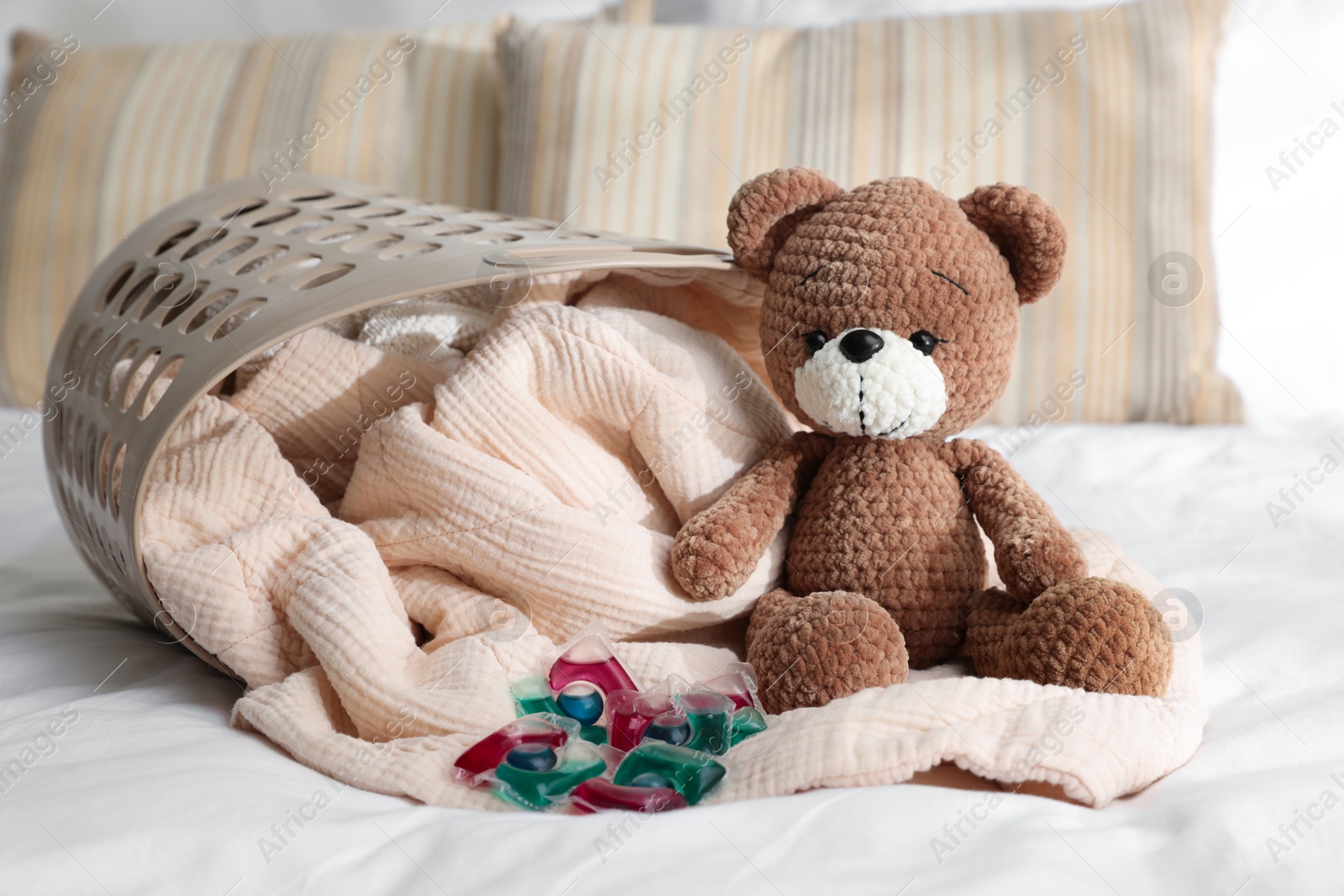 Photo of Laundry detergent capsules, teddy bear and basket with clean linens on bed indoors