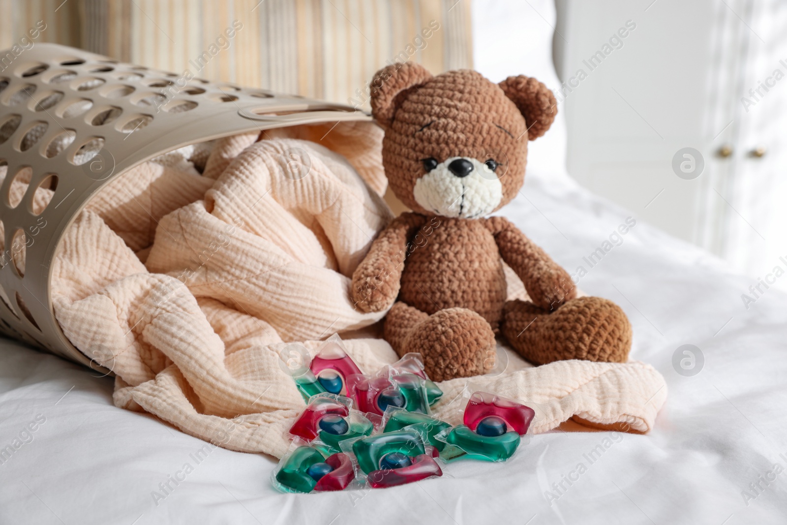 Photo of Laundry detergent capsules, teddy bear and basket with clean linens on bed indoors