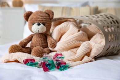 Photo of Laundry detergent capsules, teddy bear and basket with clean linens on bed indoors