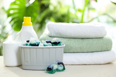 Photo of Container with laundry capsules, detergents and clean towels on table against blurred background, selective focus