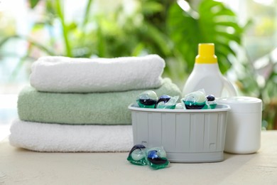 Photo of Container with laundry capsules, detergents and clean towels on table against blurred background, selective focus