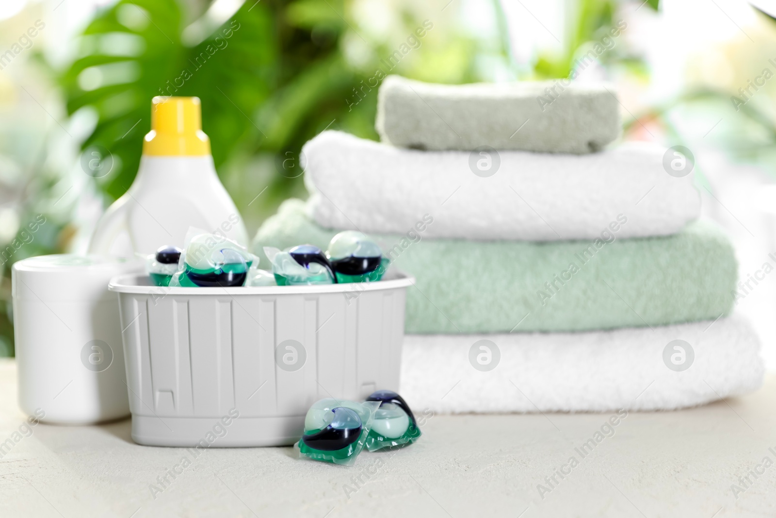 Photo of Container with laundry capsules, detergents and clean towels on table against blurred background, selective focus