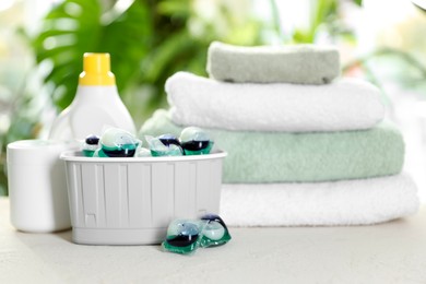 Photo of Container with laundry capsules, detergents and clean towels on table against blurred background, selective focus