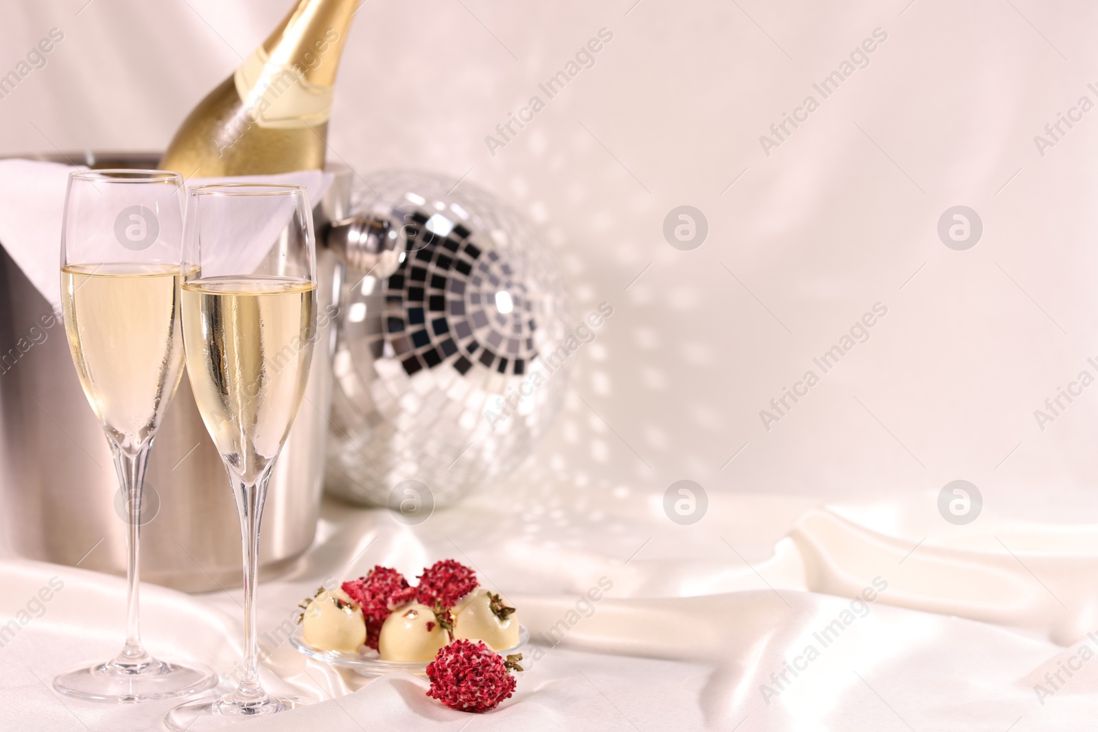 Photo of Champagne in glasses, ice bucket with bottle and tasty candies on white fabric, space for text