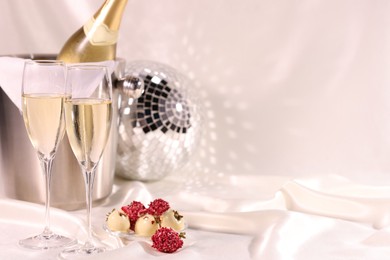 Photo of Champagne in glasses, ice bucket with bottle and tasty candies on white fabric, space for text