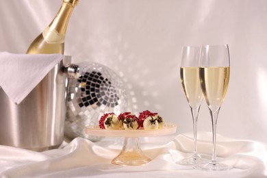 Photo of Champagne in glasses, ice bucket with bottle and tasty candies on white fabric