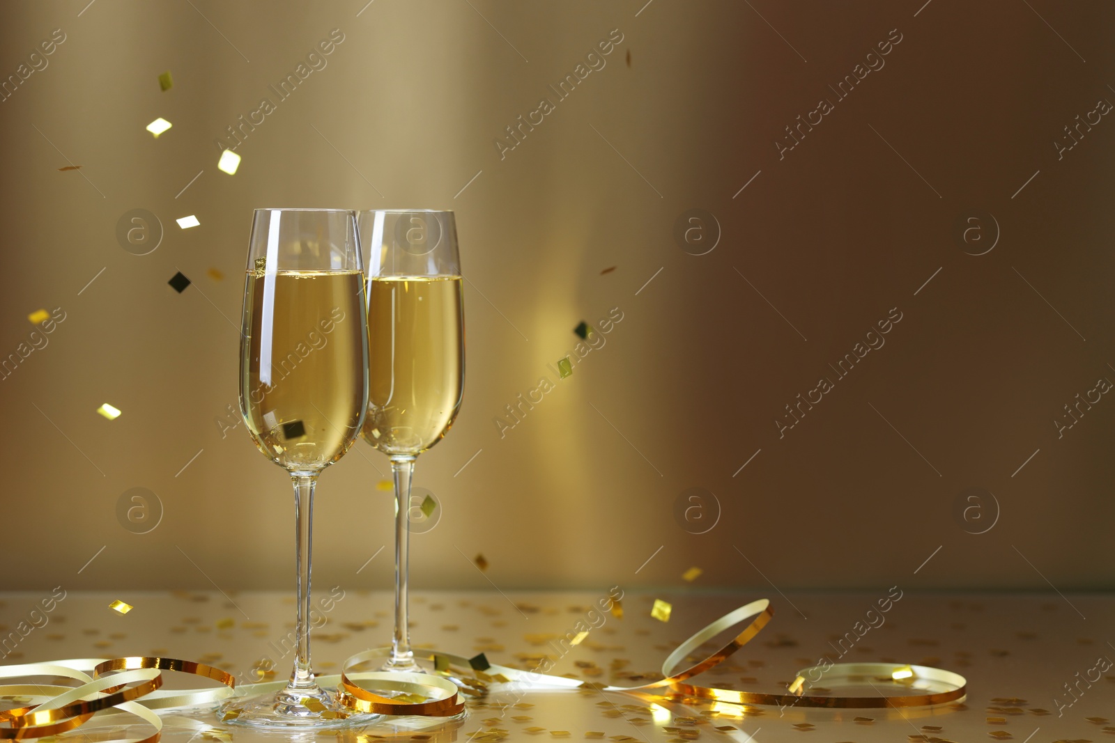 Photo of Champagne in glasses and flying confetti on golden background, space for text