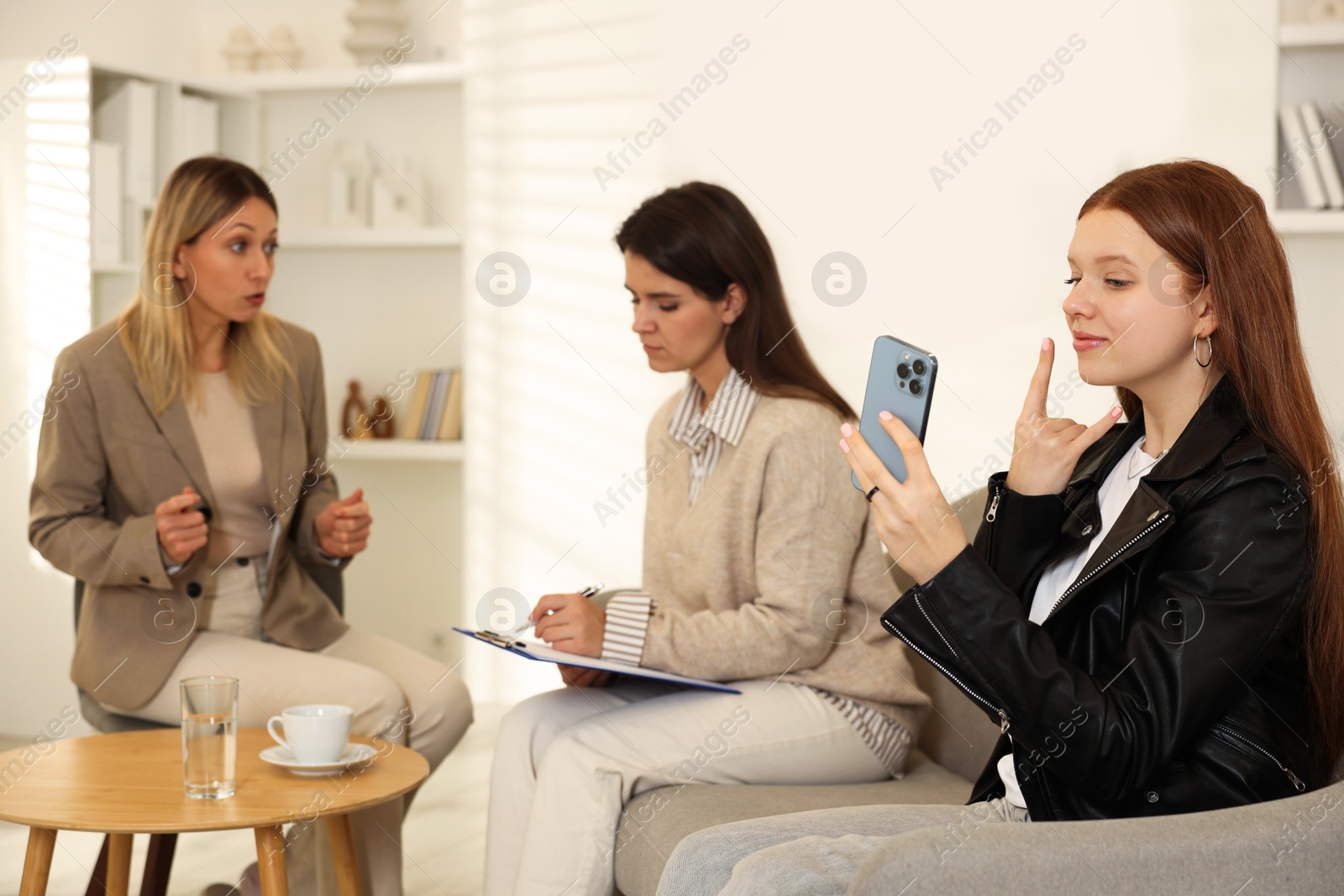 Photo of Rebellious teenage girl taking selfie during consultation with her mom and psychologist in office, selective focus