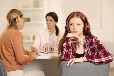 Photo of Annoyed teenage girl listening to music instead of working with psychologist in office, selective focus. Mother and daughter difficult relationship