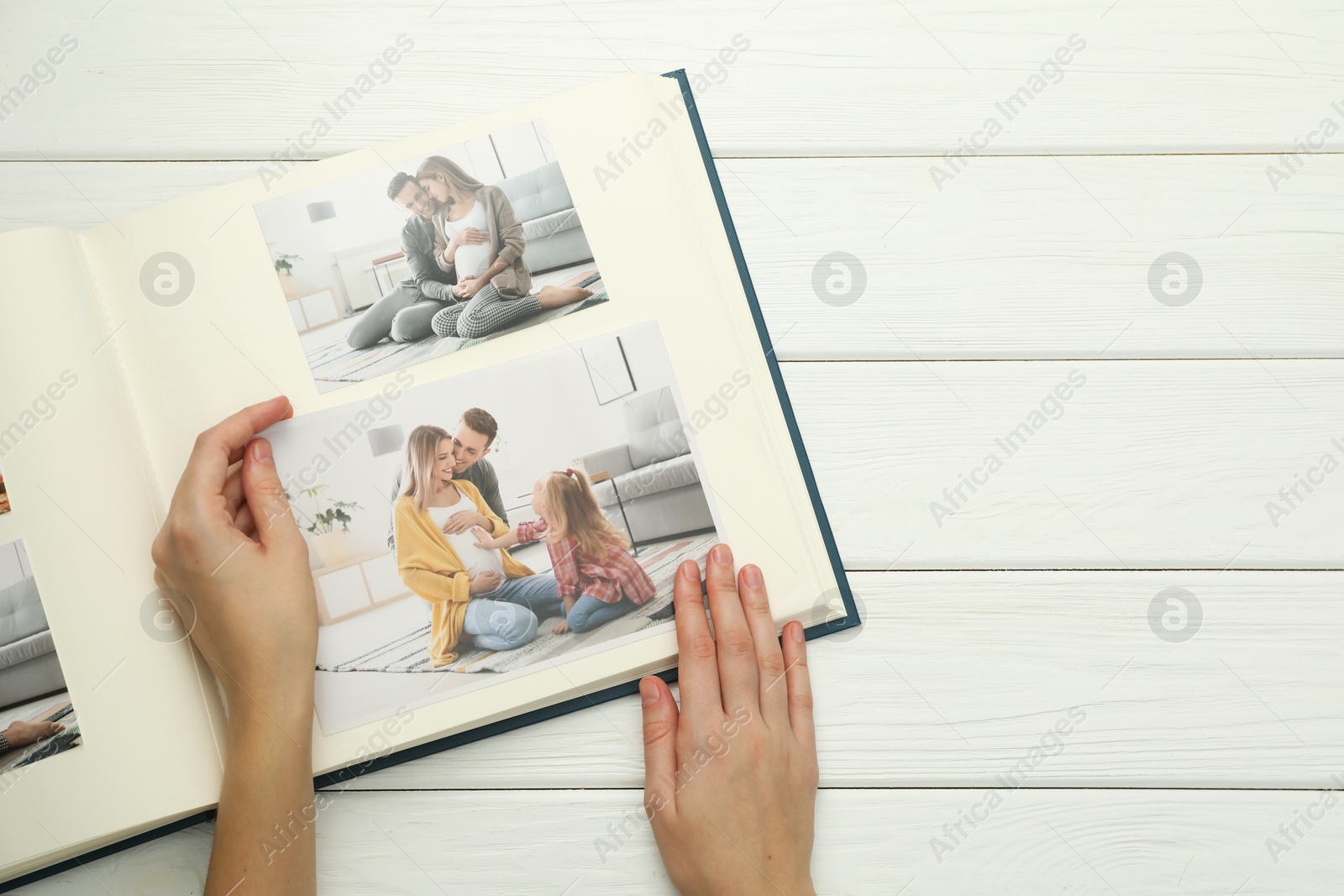 Photo of Woman with different photos in open photo album at white wooden table, top view. Space for text