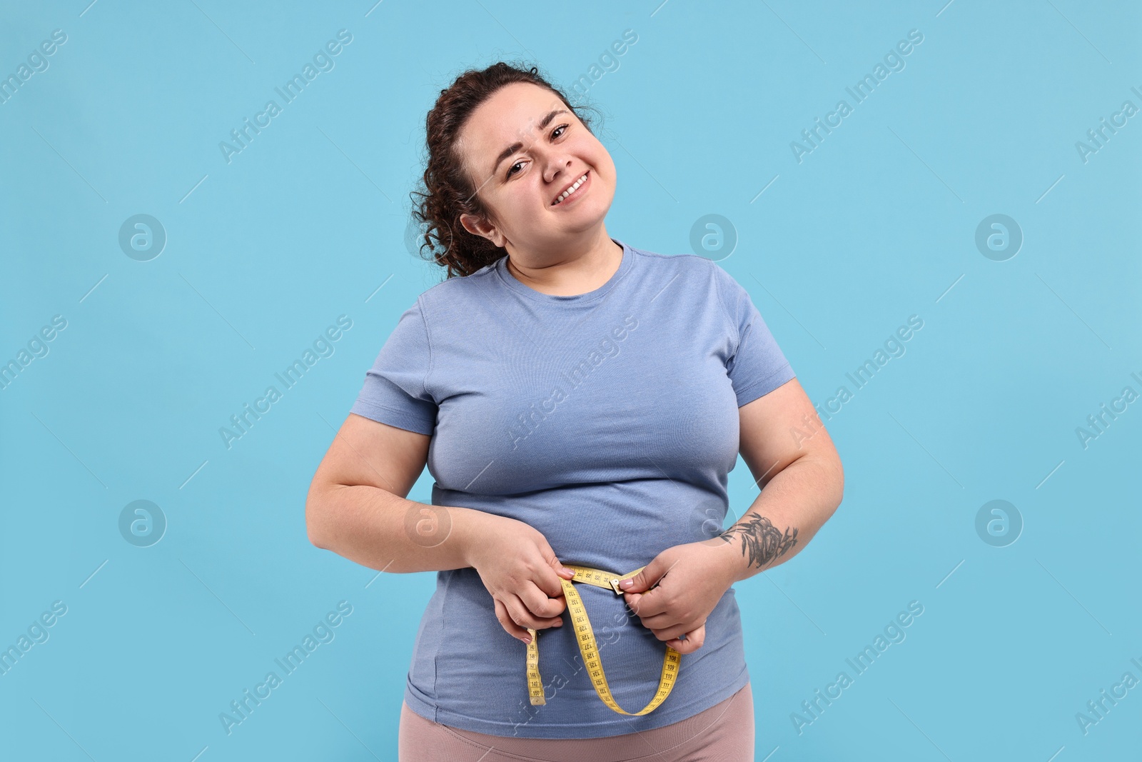 Photo of Weight loss. Happy plus size woman measuring waist with tape on light blue background