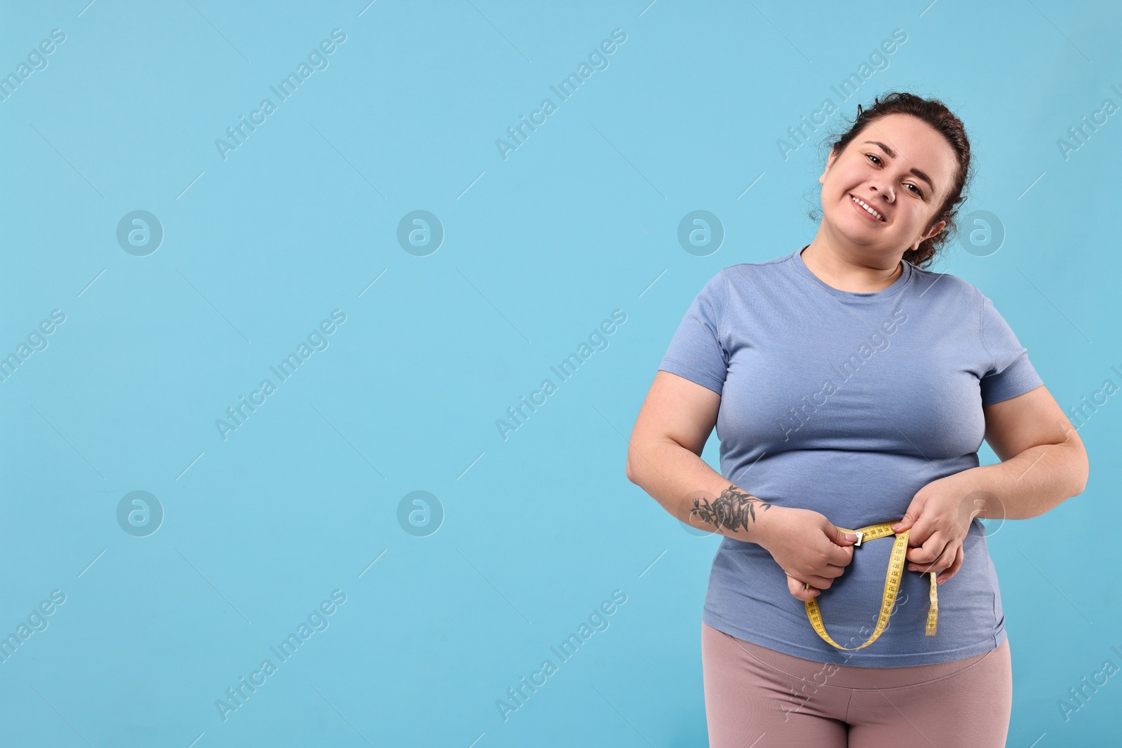 Photo of Weight loss. Happy plus size woman measuring waist with tape on light blue background, space for text