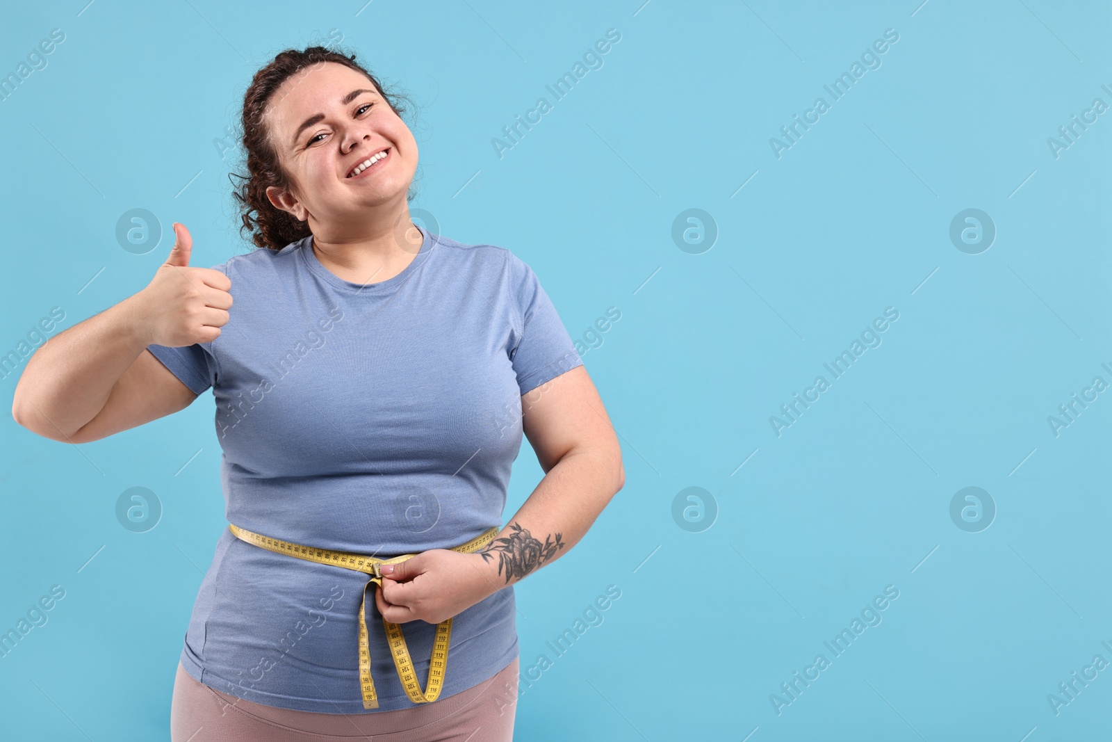 Photo of Weight loss. Happy plus size woman measuring waist with tape and showing thumbs up on light blue background, space for text