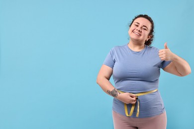 Weight loss. Happy plus size woman measuring waist with tape and showing thumbs up on light blue background, space for text