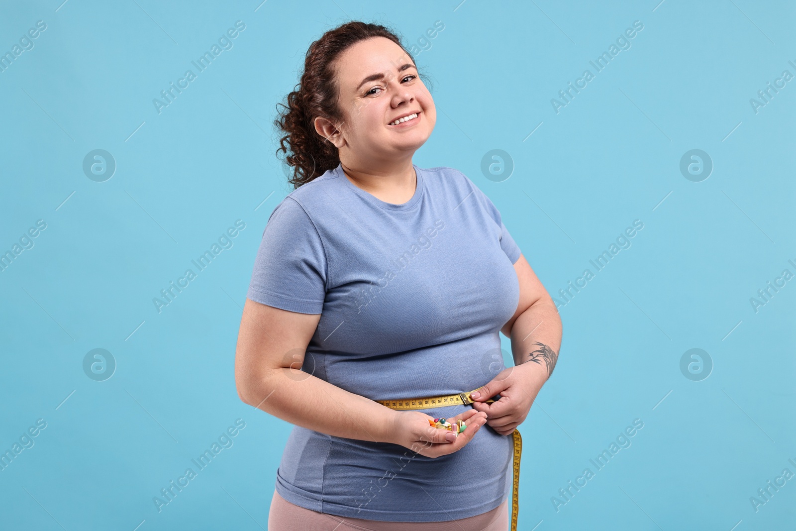 Photo of Happy plus size woman measuring waist with tape and holding pile of weight loss supplements on light blue background