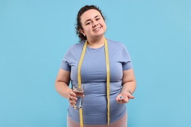 Photo of Happy plus size woman holding pile of weight loss supplements and glass with water on light blue background