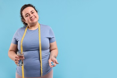 Happy plus size woman holding pile of weight loss supplements and glass with water on light blue background. Space for text