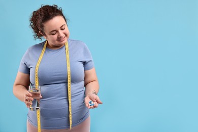 Photo of Happy plus size woman holding pile of weight loss supplements and glass with water on light blue background. Space for text