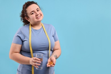 Photo of Happy plus size woman holding pile of weight loss supplements and glass with water on light blue background. Space for text