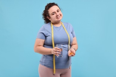 Photo of Happy plus size woman holding pile of weight loss supplements and glass with water on light blue background