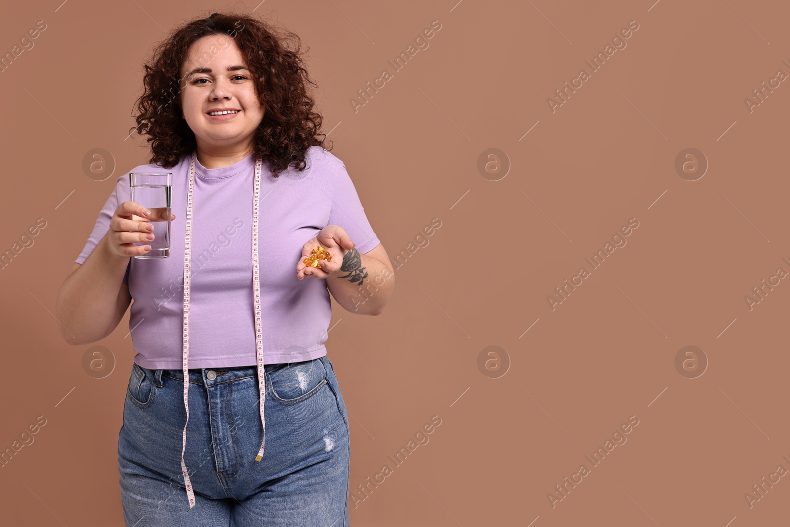 Photo of Happy plus size woman holding pile of weight loss supplements and glass of water on beige background. Space for text