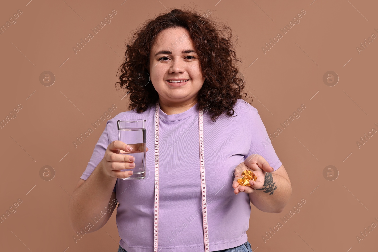 Photo of Happy plus size woman holding pile of weight loss supplements and glass of water on beige background