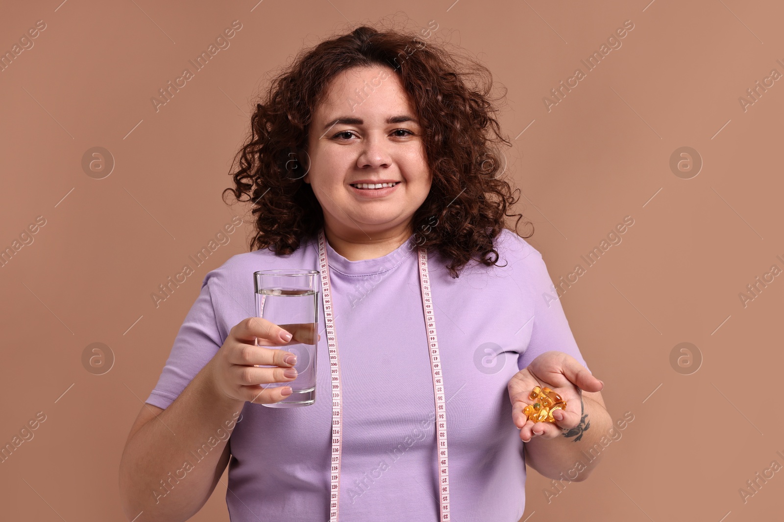 Photo of Happy plus size woman holding pile of weight loss supplements and glass of water on beige background