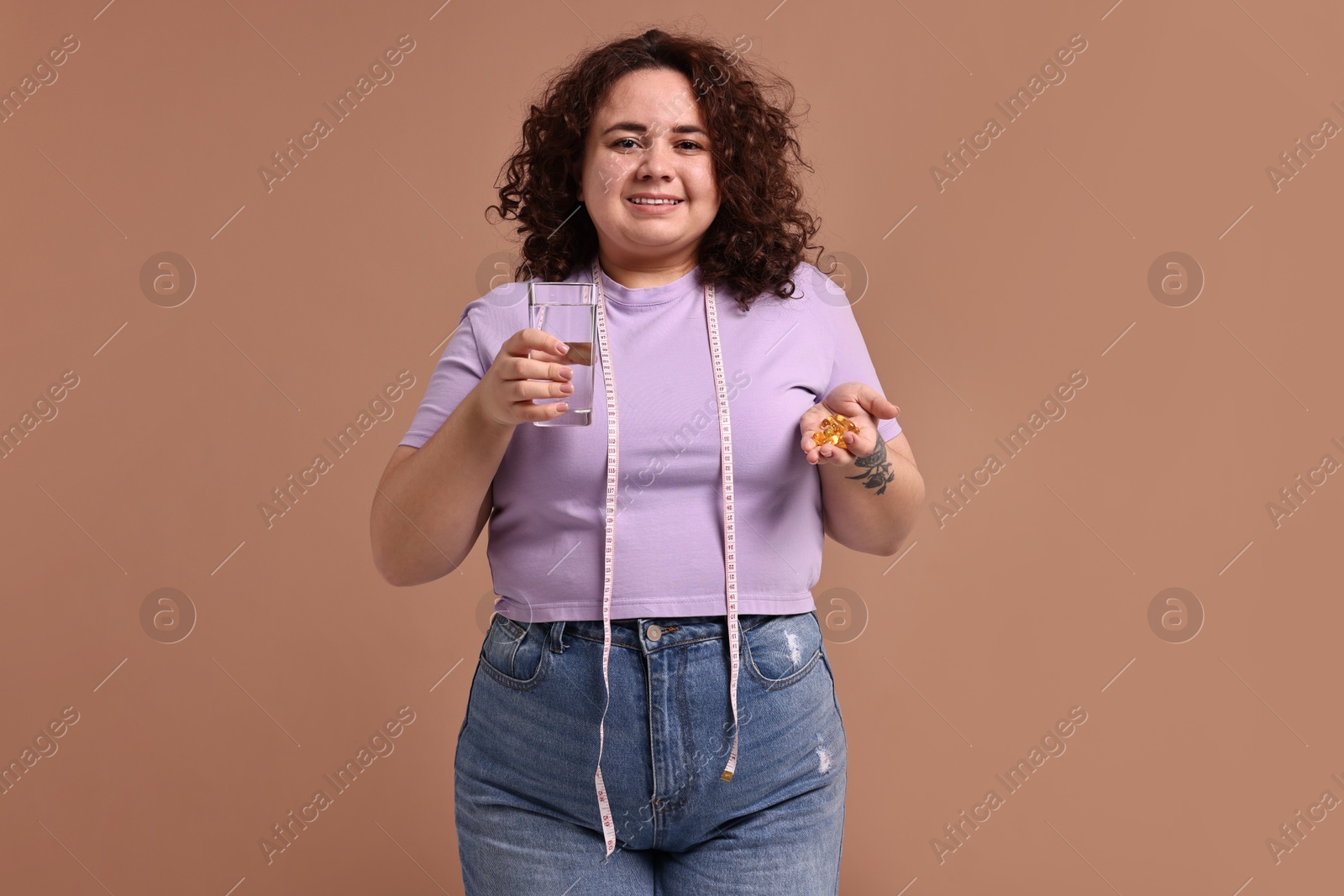 Photo of Happy plus size woman holding pile of weight loss supplements and glass of water on beige background