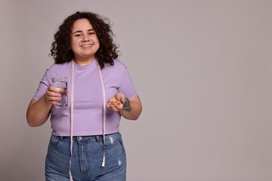 Photo of Happy plus size woman holding pile of weight loss supplements and glass of water on grey background. Space for text