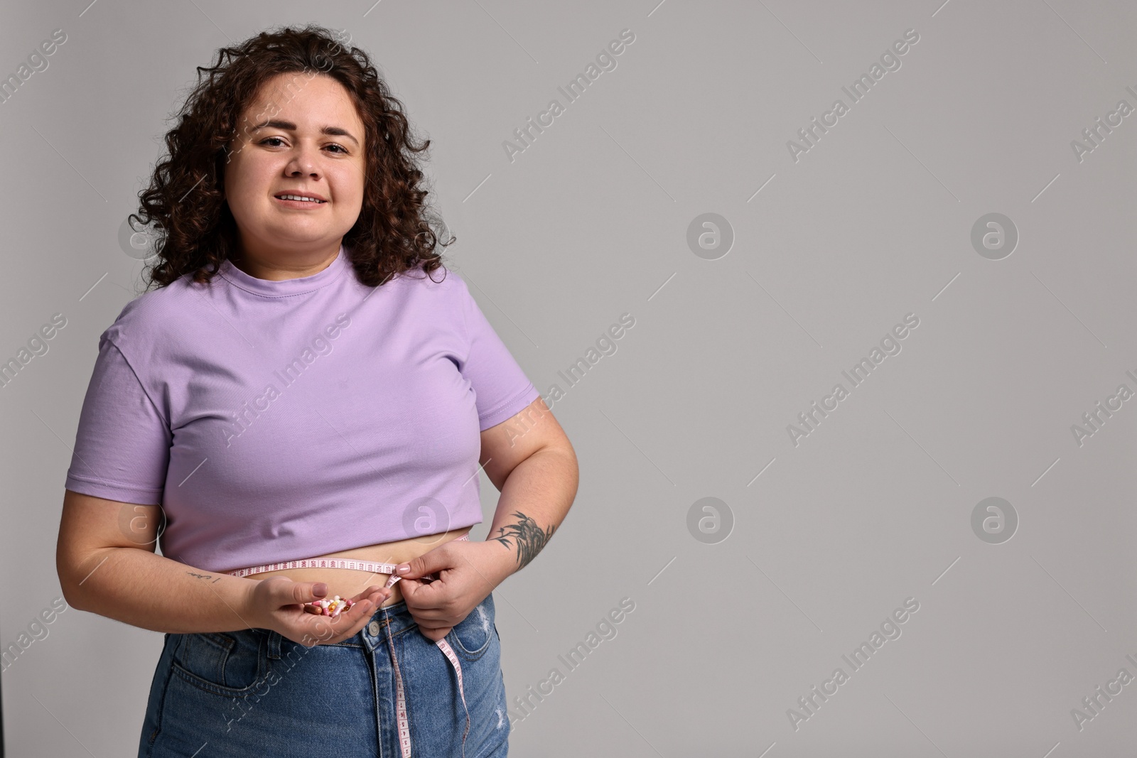 Photo of Happy plus size woman measuring waist with tape and holding pile of weight loss supplements on grey background. Space for text