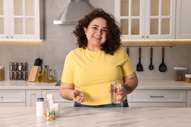 Photo of Happy plus size woman with weight loss supplements and glass of water at marble countertop in kitchen