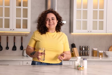 Happy plus size woman with weight loss supplements and glass of water at marble countertop in kitchen