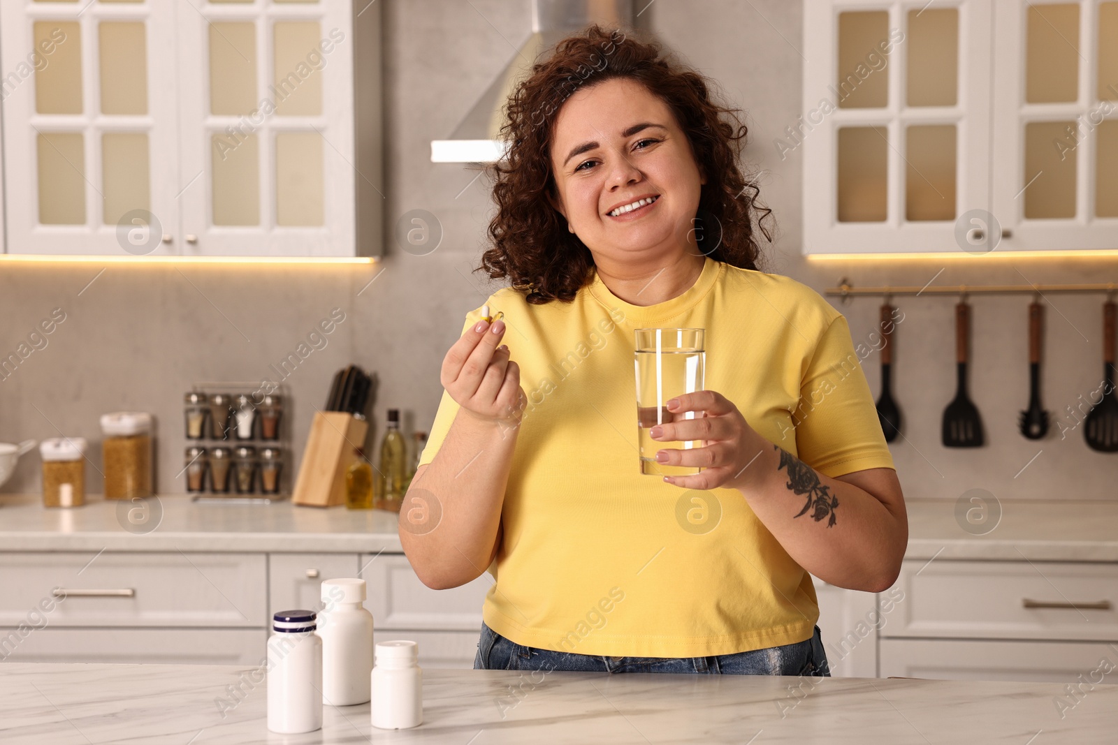 Photo of Happy plus size woman with weight loss supplements and glass of water at marble countertop in kitchen