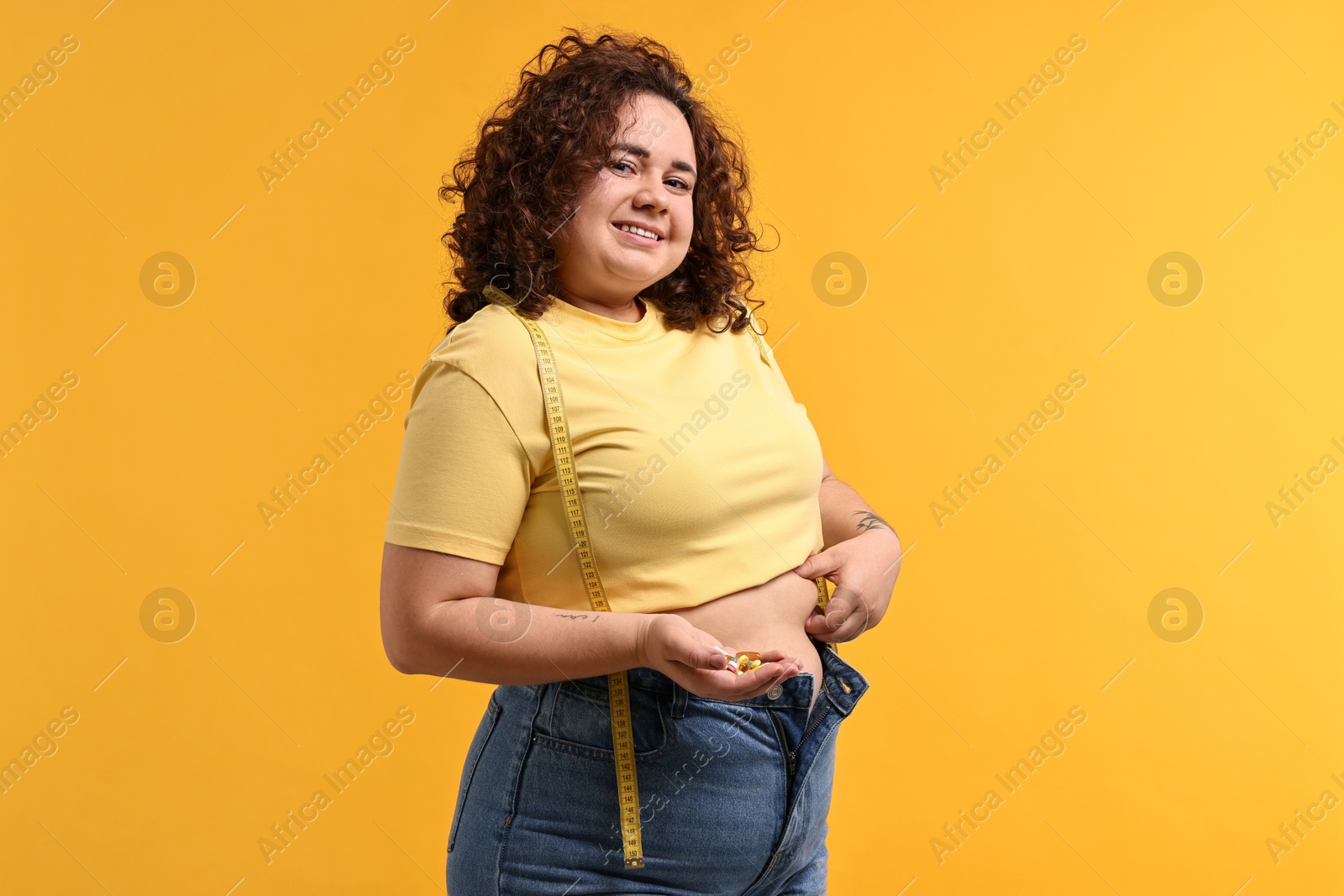 Photo of Happy plus size woman with pile of weight loss supplements on orange background